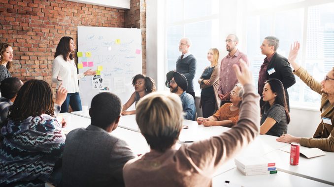 Diverse group of people in a meeting around a table and standing, as female leader presents.